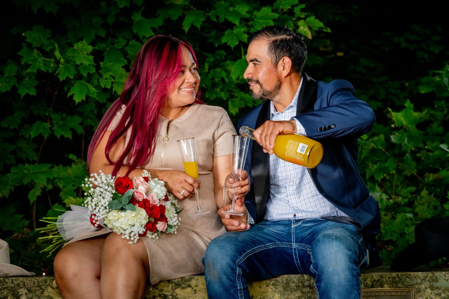 Happy couple celebrating with champagne and wedding bouquet seated outdoors among green foliage.