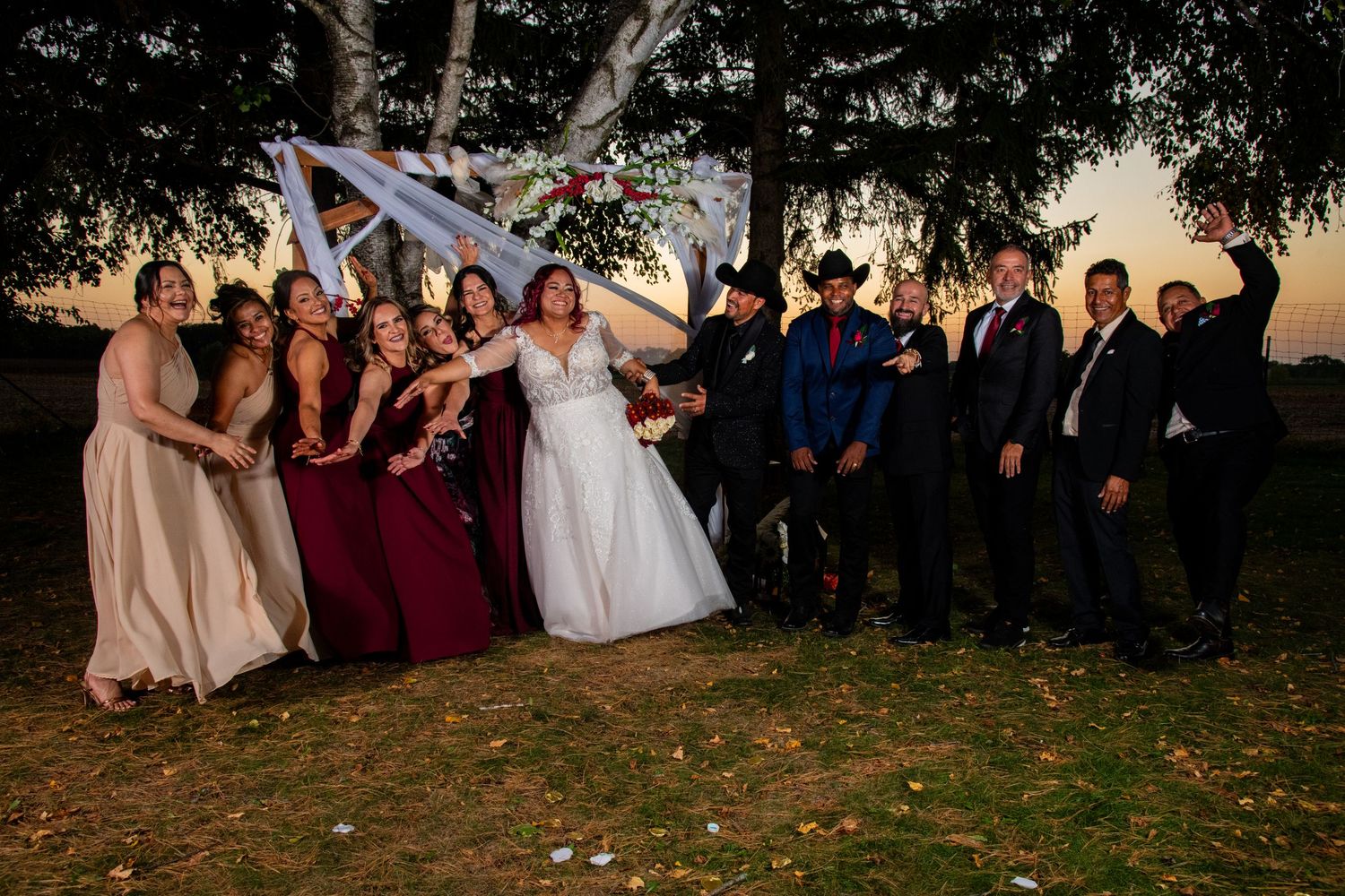 Large wedding party group photo at sunset under decorated arch with bridesmaids and groomsmen.