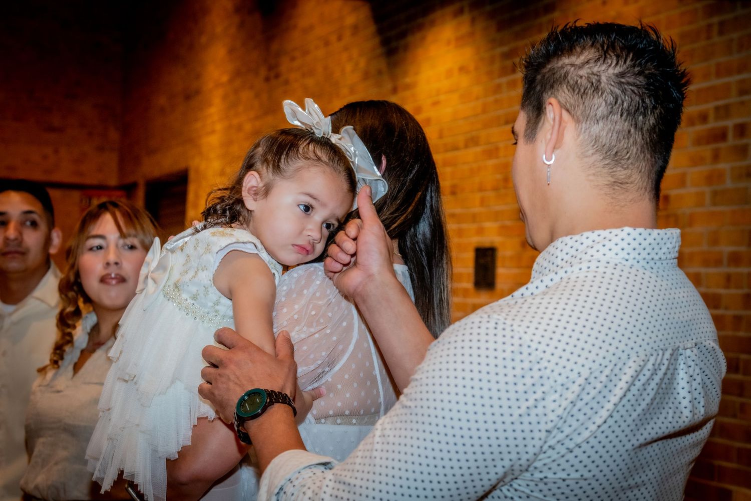 A man gently touches a little girl in a white dress held by a woman at a family celebration near a brick wall.