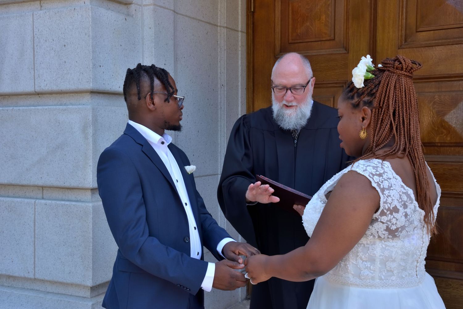 Bride and groom exchange vows before bearded officiant on courthouse steps in civil ceremony.
