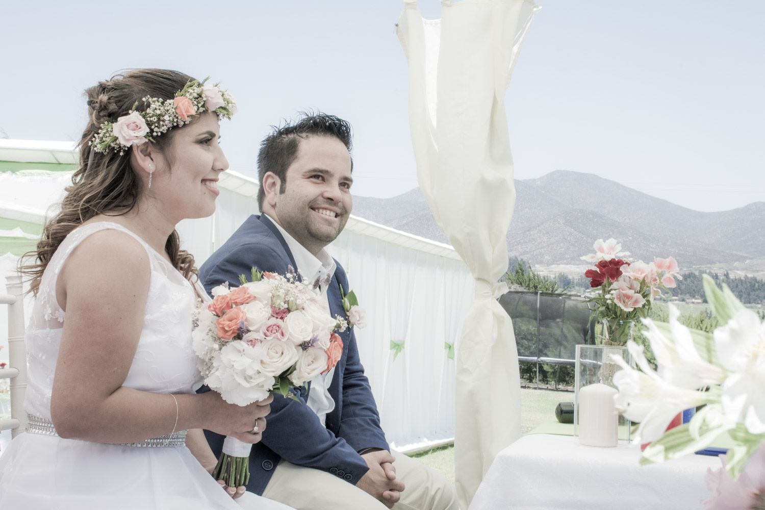Bride with floral crown holding pink bouquet sits beside groom in blue suit during outdoor wedding ceremony.