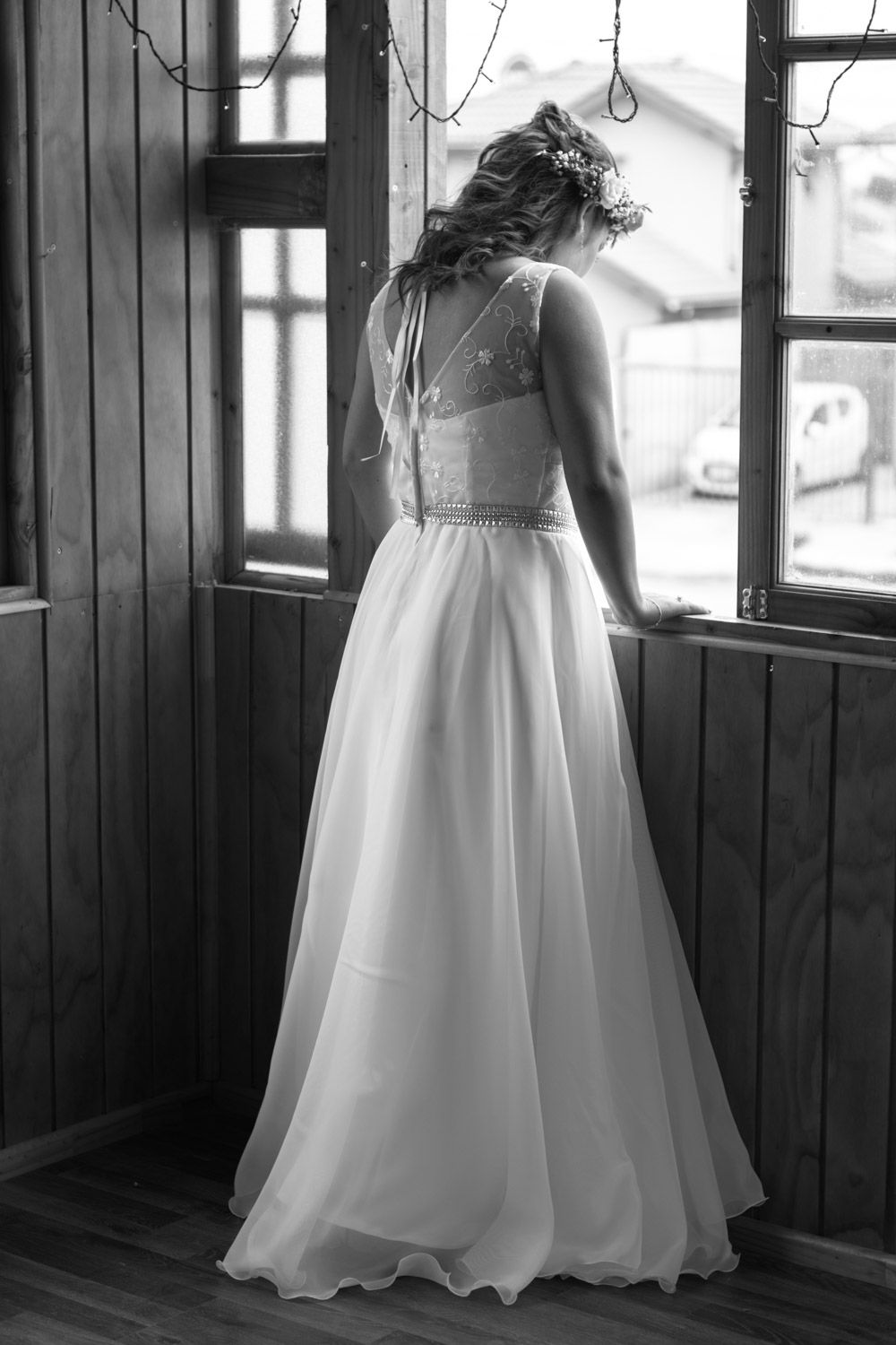 Bride in elegant white lace wedding gown viewed from behind standing by wooden window in black and white photo.