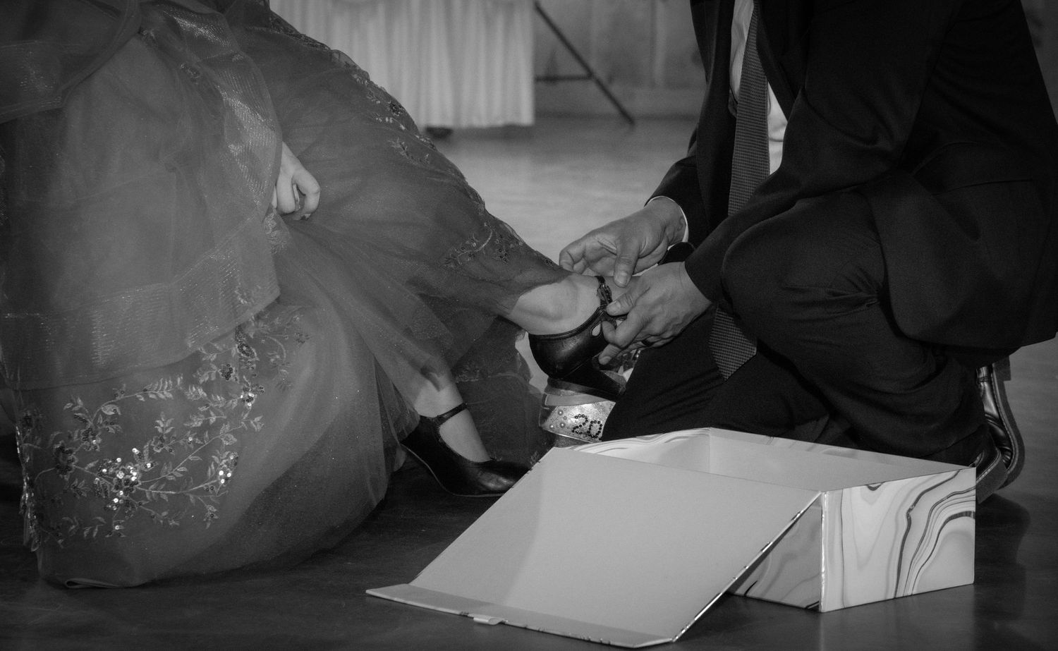 Black and white photo of a groom helping bride with her shoe during wedding reception on floor.