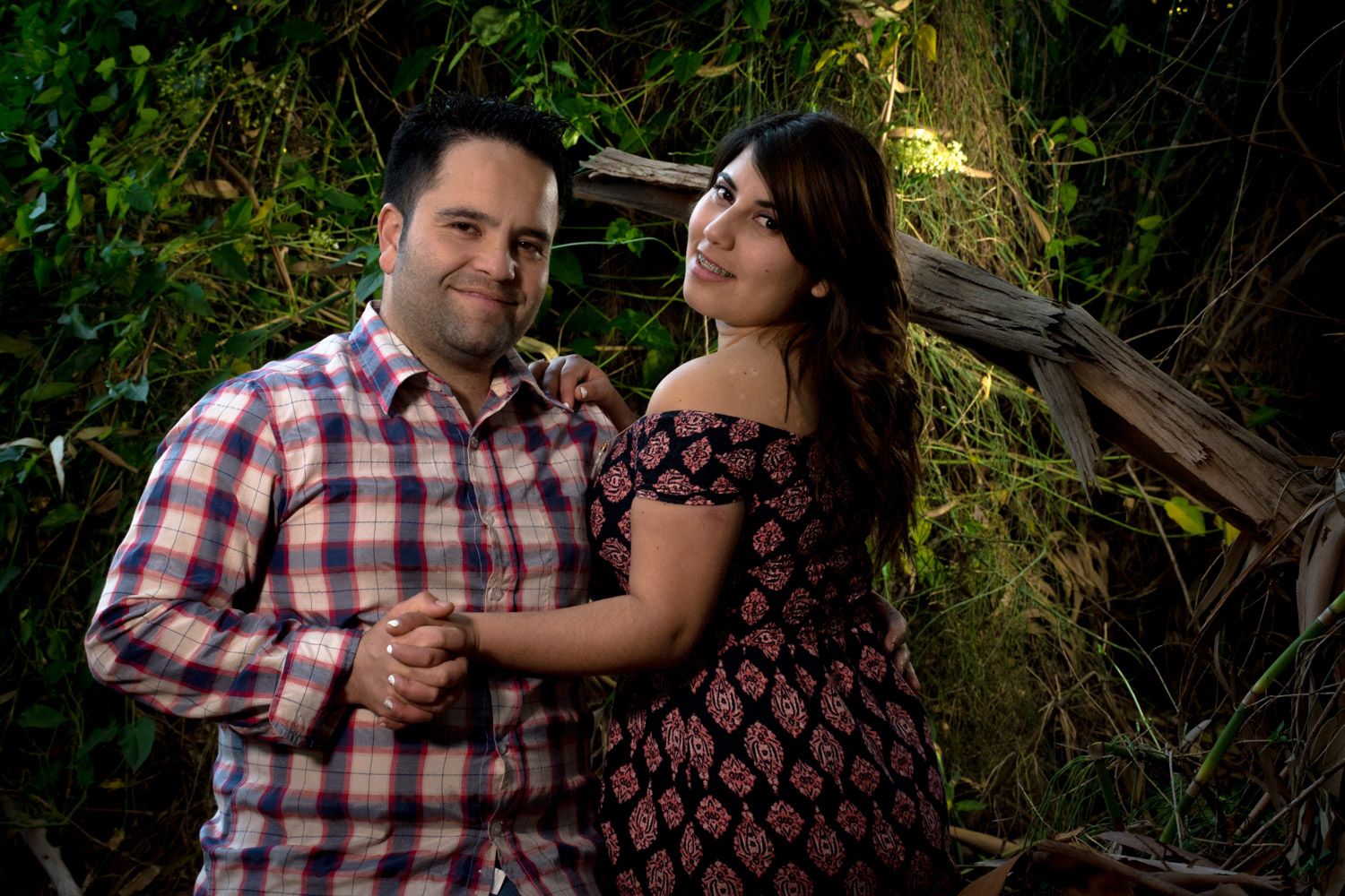 Smiling couple holding hands while posing against lush green ivy-covered wall outdoors.