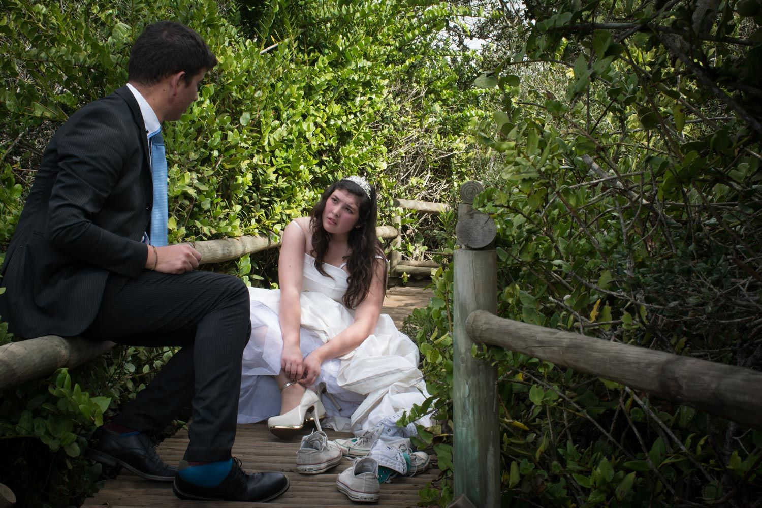 Bride and groom sit together on a wooden bridge surrounded by lush green foliage.