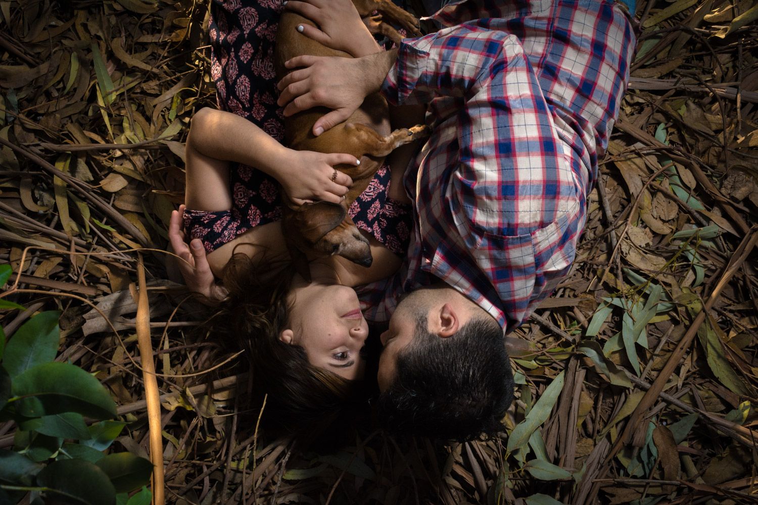 Happy couple laughing together while lying on forest floor surrounded by green leaves.
