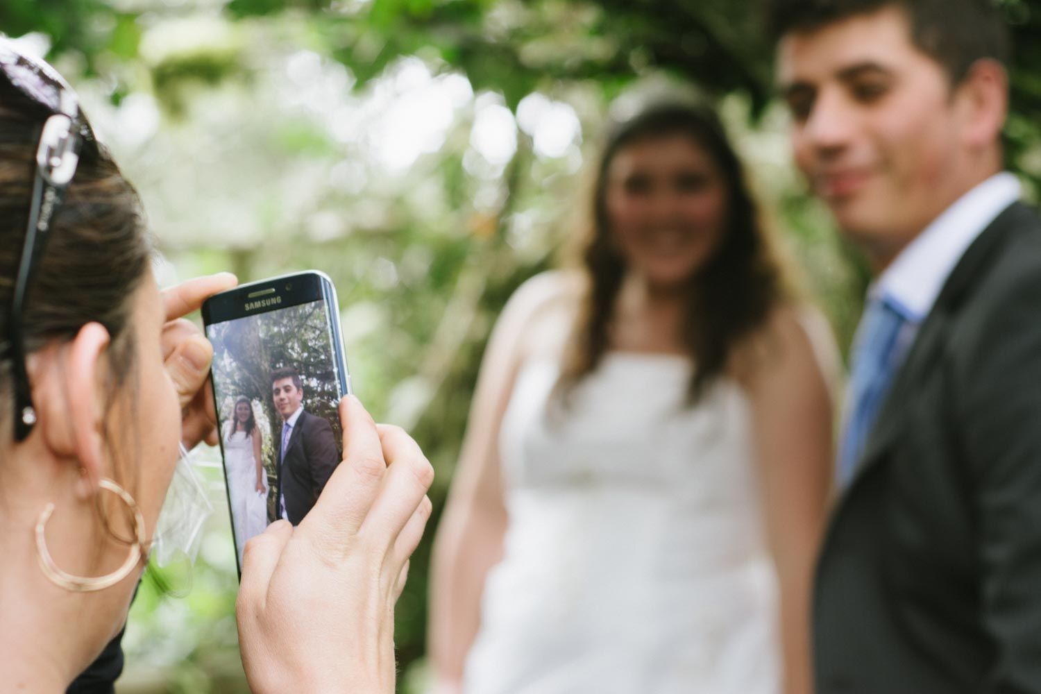 Guest photographing smiling bride and groom on smartphone at outdoor wedding celebration.