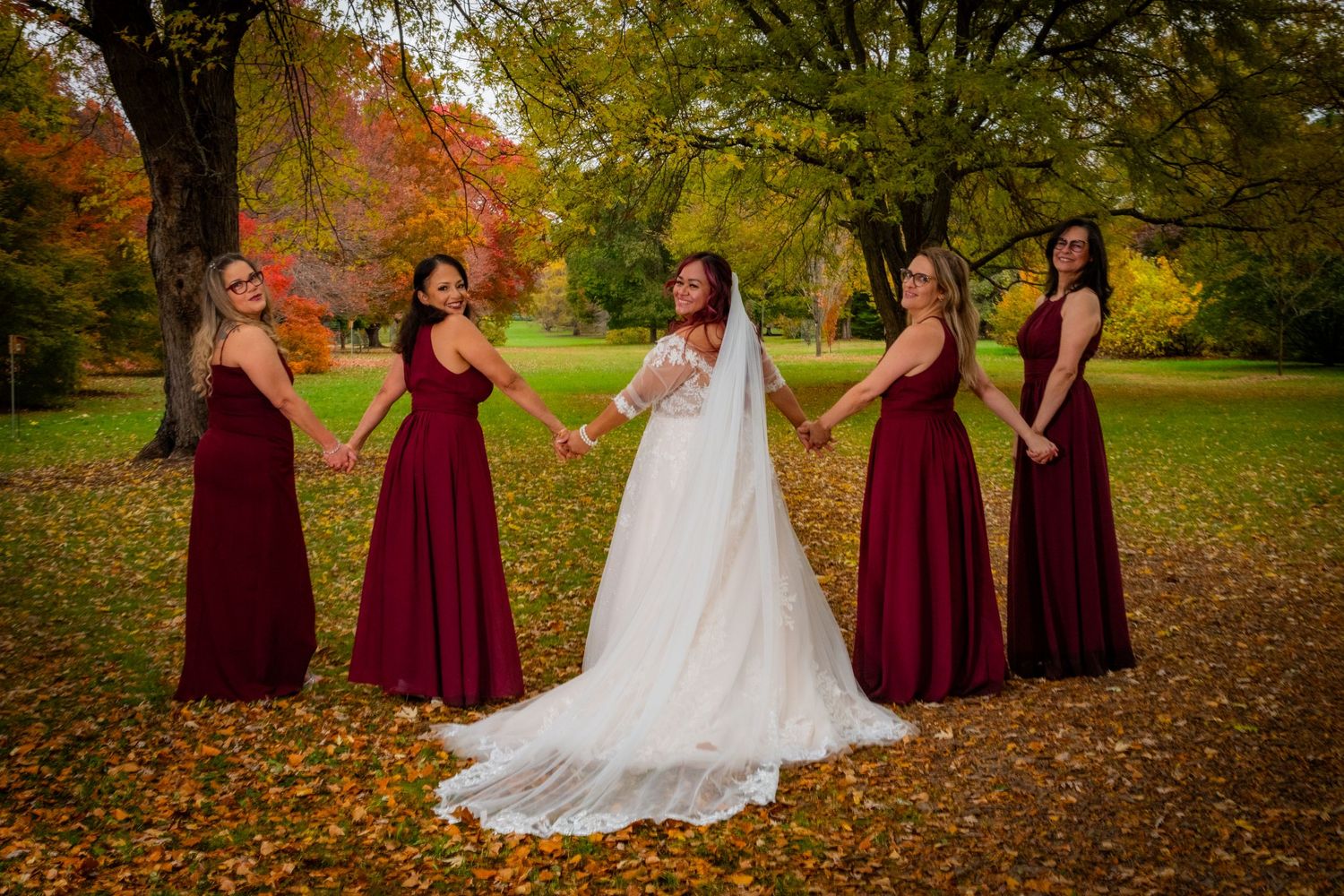 Bride and four bridesmaids in burgundy gowns hold hands in a colorful autumn park setting.