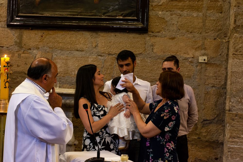 Priest performing a baptism ceremony with family gathered around a baby in a white christening gown inside a stone church.