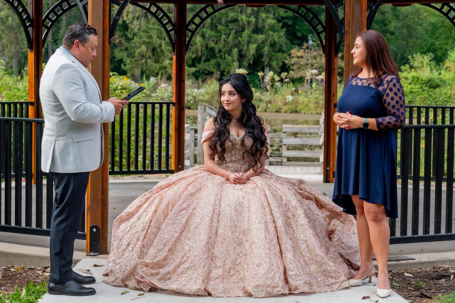 Quinceañera ceremony in gazebo with girl in rose gold gown receiving blessing from officiants outdoors.
