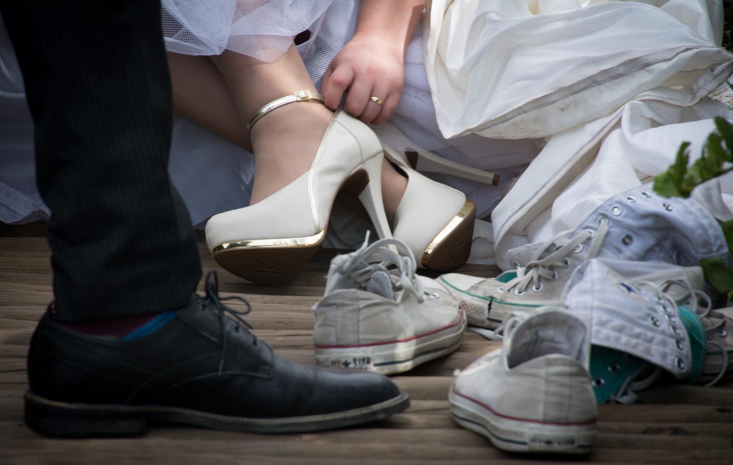 Close-up of bride adjusting white heels beside groom wearing casual sneakers on wooden floor.