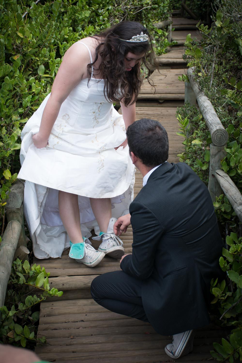 Groom kneeling on wooden boardwalk helping bride with sneakers under wedding dress.
