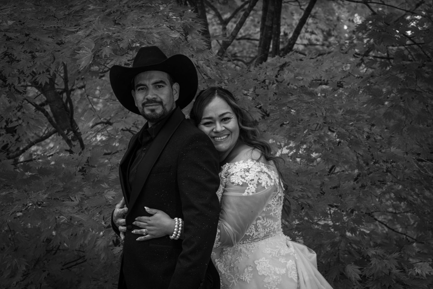 Black and white photo of a bride and groom posing together outdoors among autumn trees.