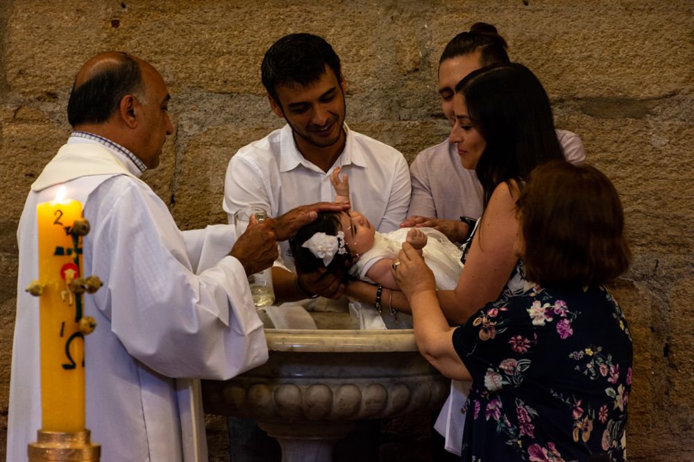Catholic priest baptizes infant at stone font surrounded by godparents in traditional church ceremony.