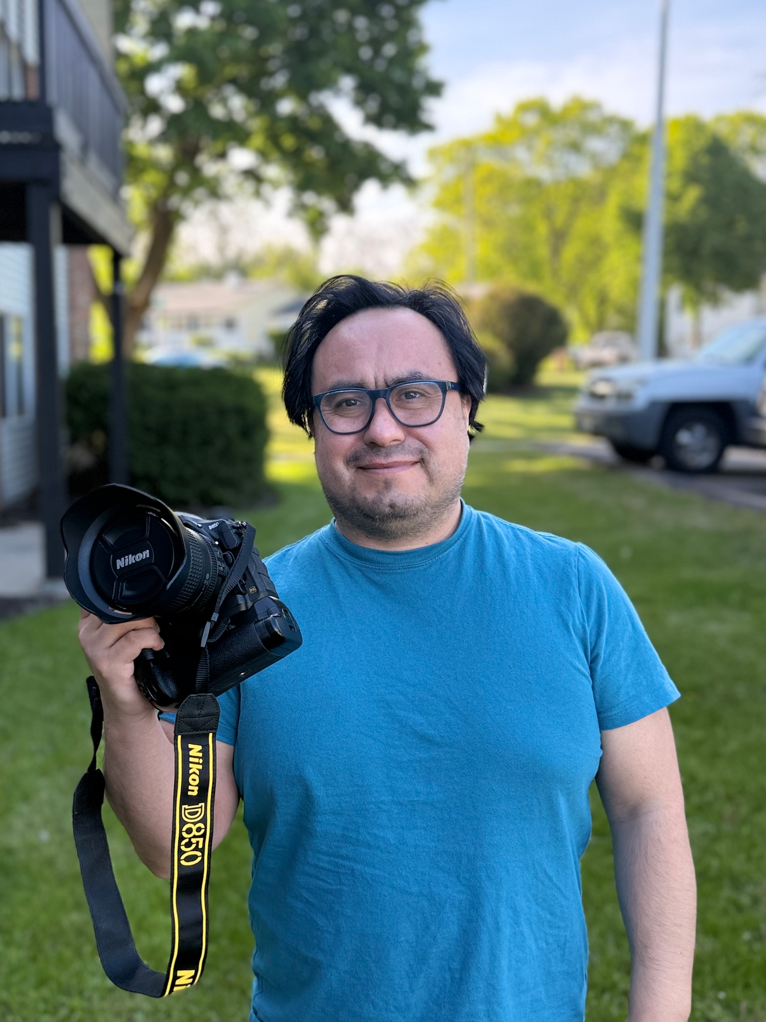 Luis holding a Nikon D850 camera outdoors, smiling, wearing glasses, with green trees in background.