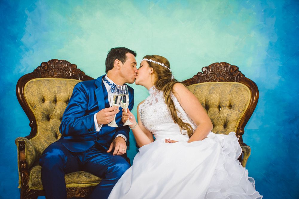 Bride and groom kissing on vintage gold sofa holding champagne glasses in front of blue backdrop.