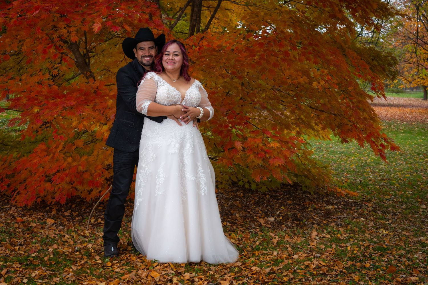 Couple poses in autumn foliage, bride in white lace gown and groom in black suit with cowboy hat.
