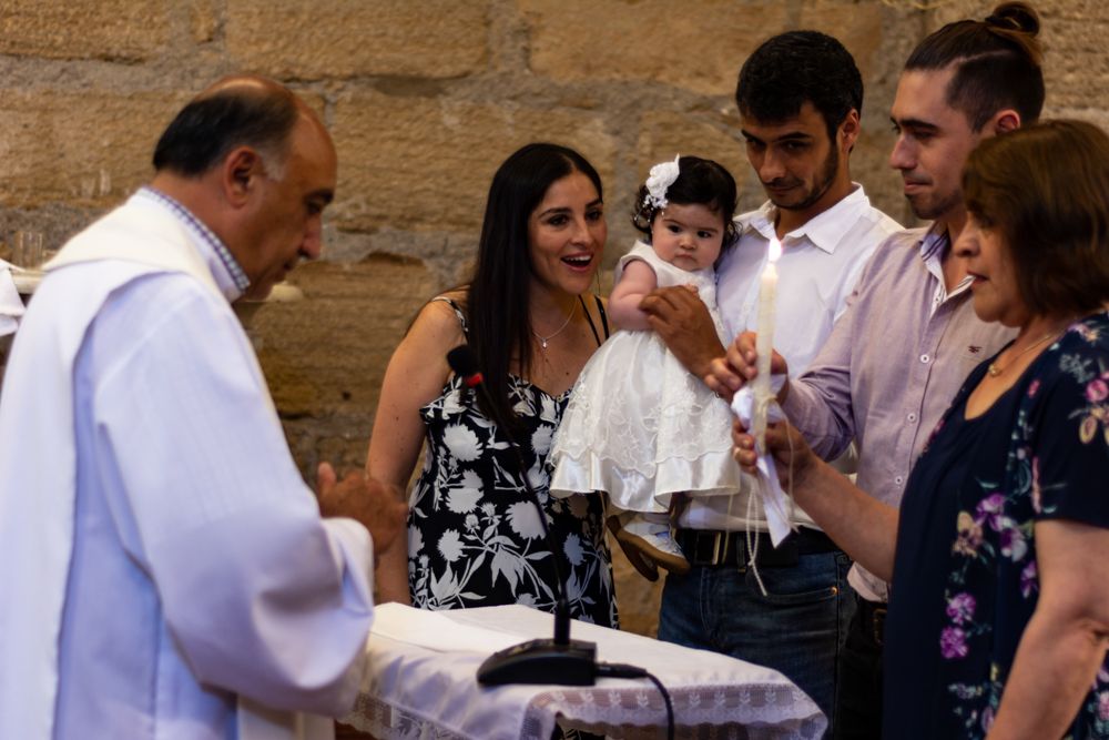 Priest performs baptism ceremony with family holding baby in white christening gown at church font.