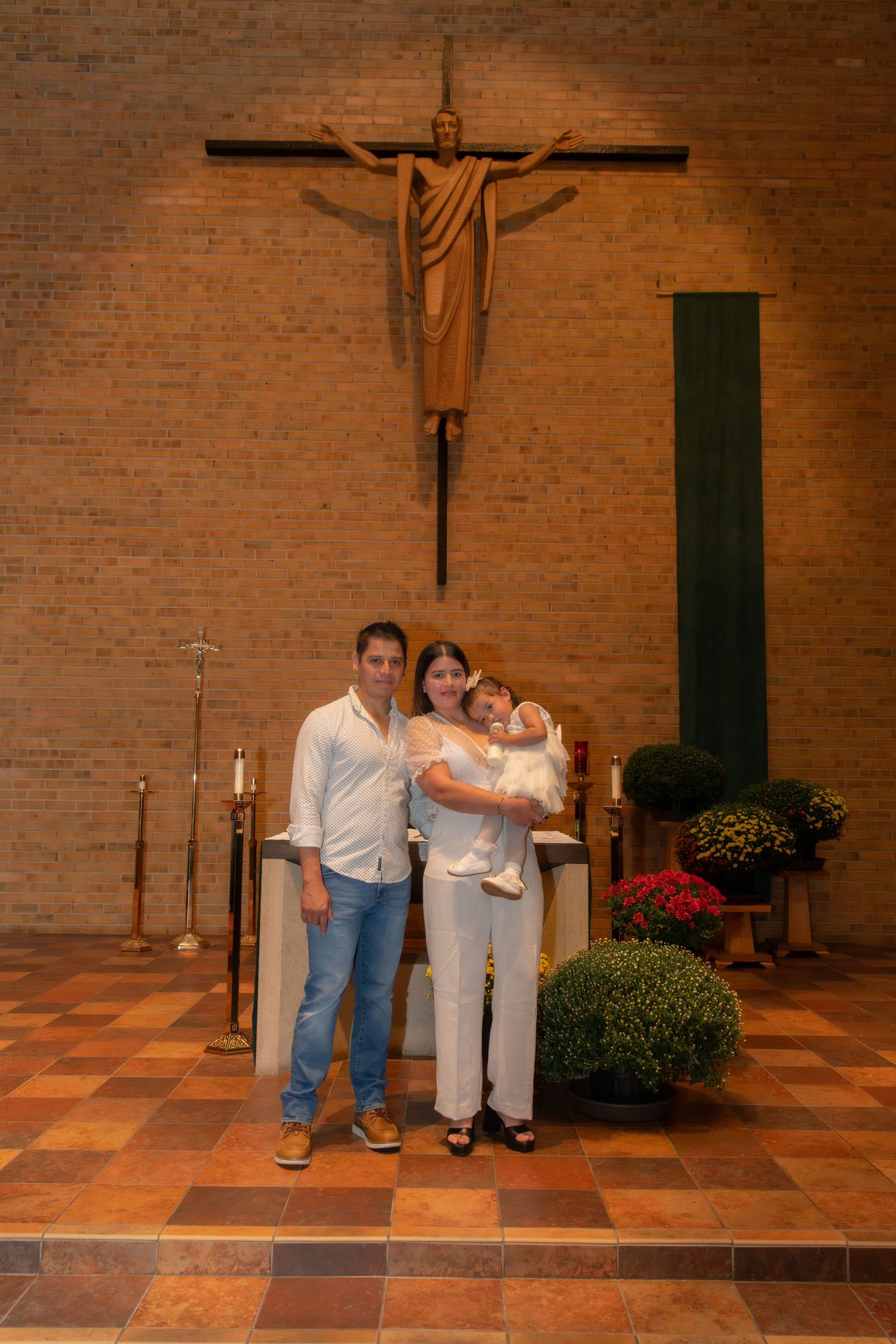 Family posing with baby in white outfit at church altar with crucifix during baptism ceremony.