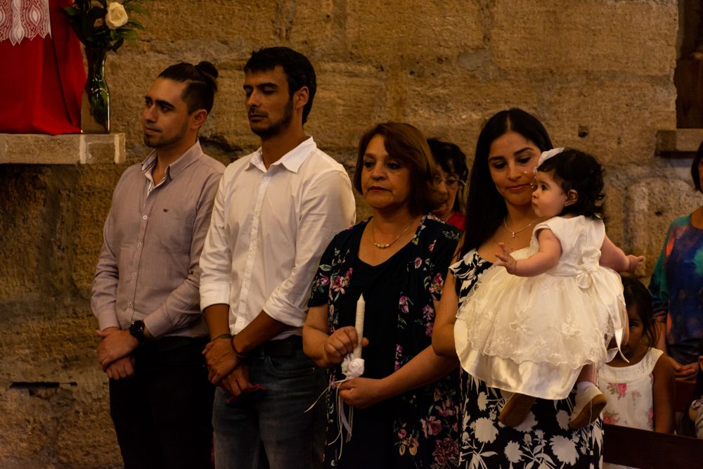 Family gathered in a church for a baptism ceremony, with a woman holding a baby in a white christening gown.