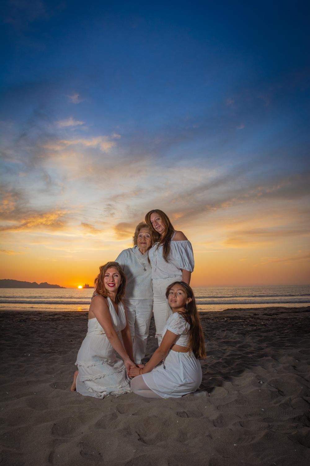Four women in white dresses pose together on sandy beach during golden sunset with vibrant sky.