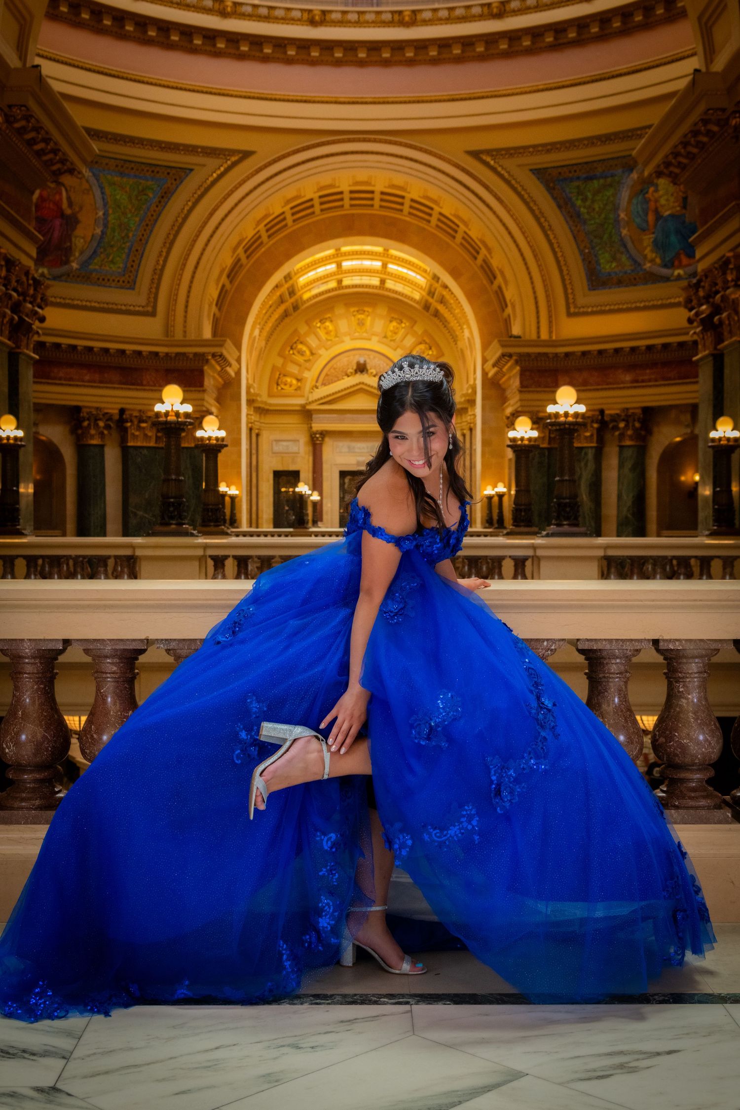 Young woman in royal blue quinceañera ball gown with tiara posing in ornate capitol building.
