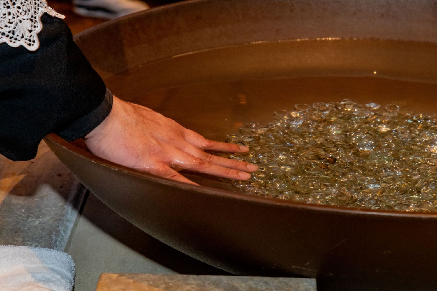 A hand touching holy water with coins in a large brown baptismal font during a religious ceremony.