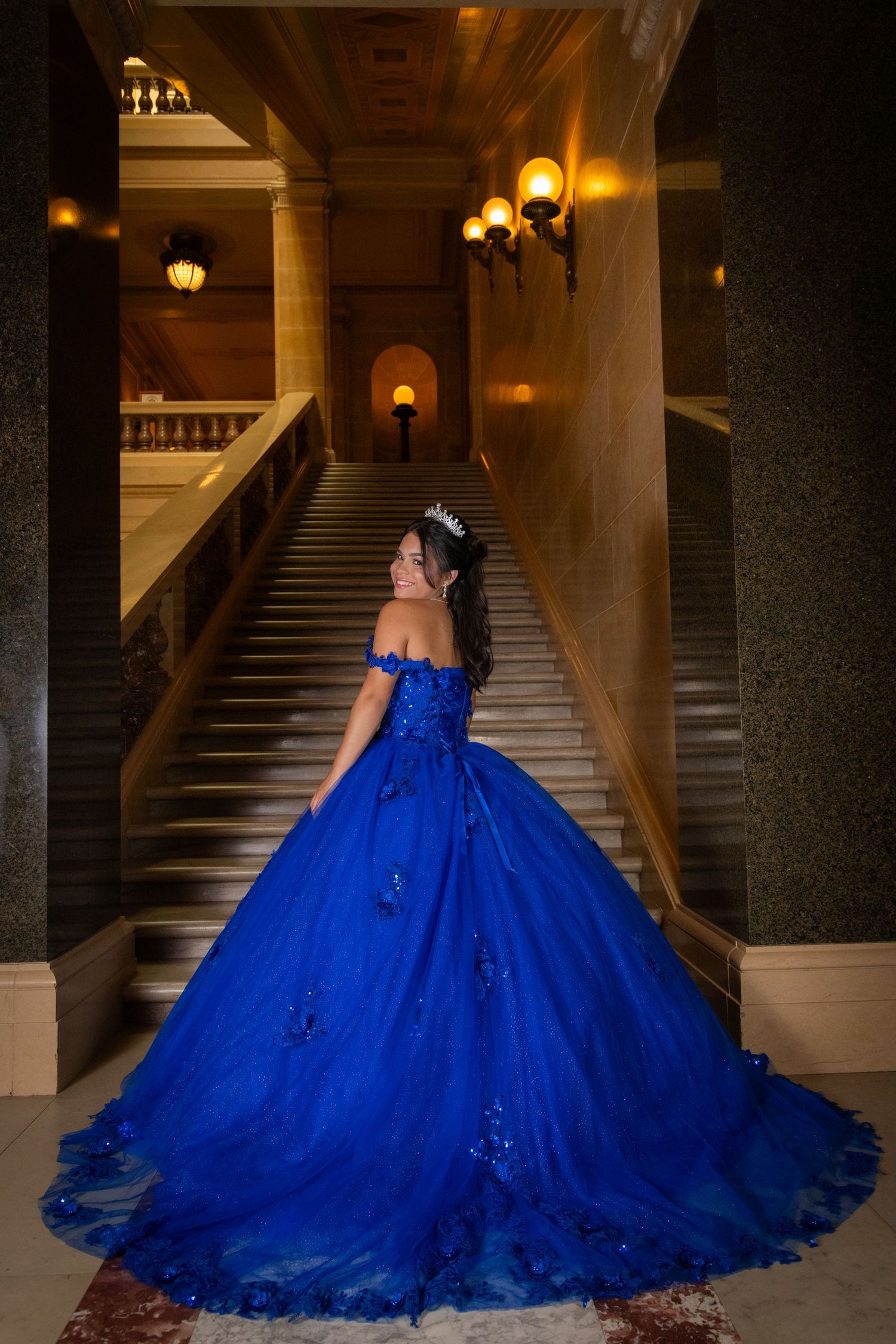 A quinceañera girl in a royal blue ball gown and tiara poses on grand marble stairs in an elegant venue.