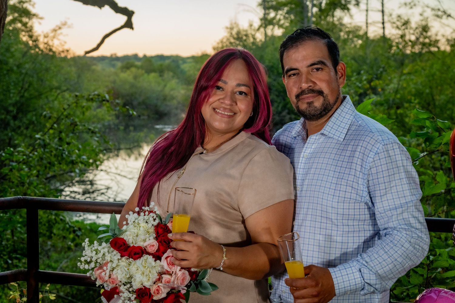 Smiling couple holding champagne glasses with a scenic river and sunset backdrop during engagement.