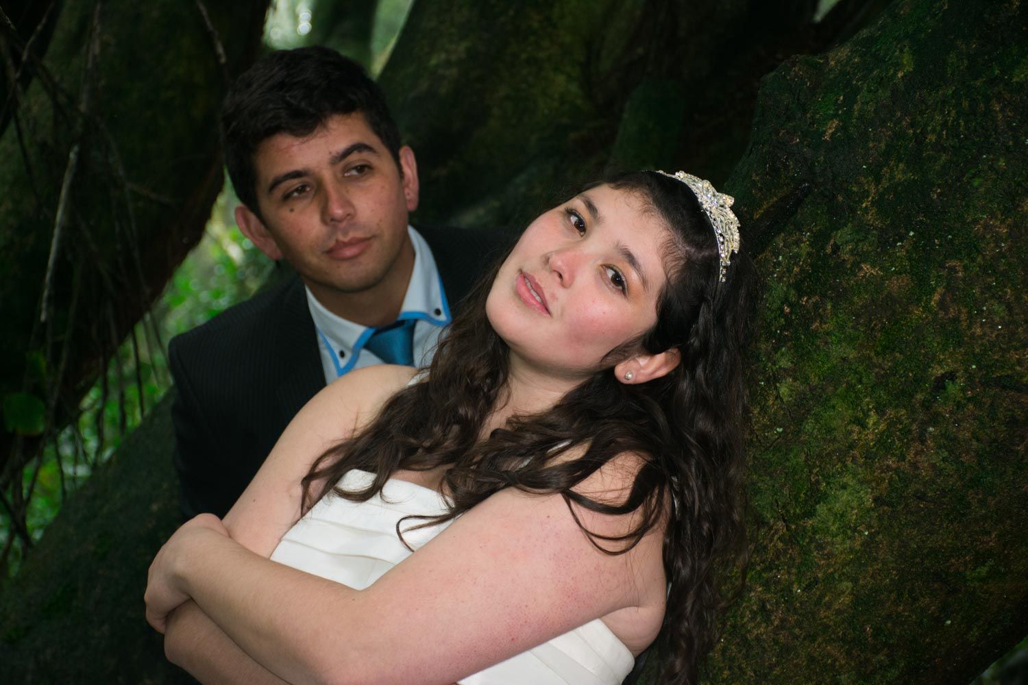 Bride in white gown with tiara leaning against groom near a moss-covered tree in a forest.