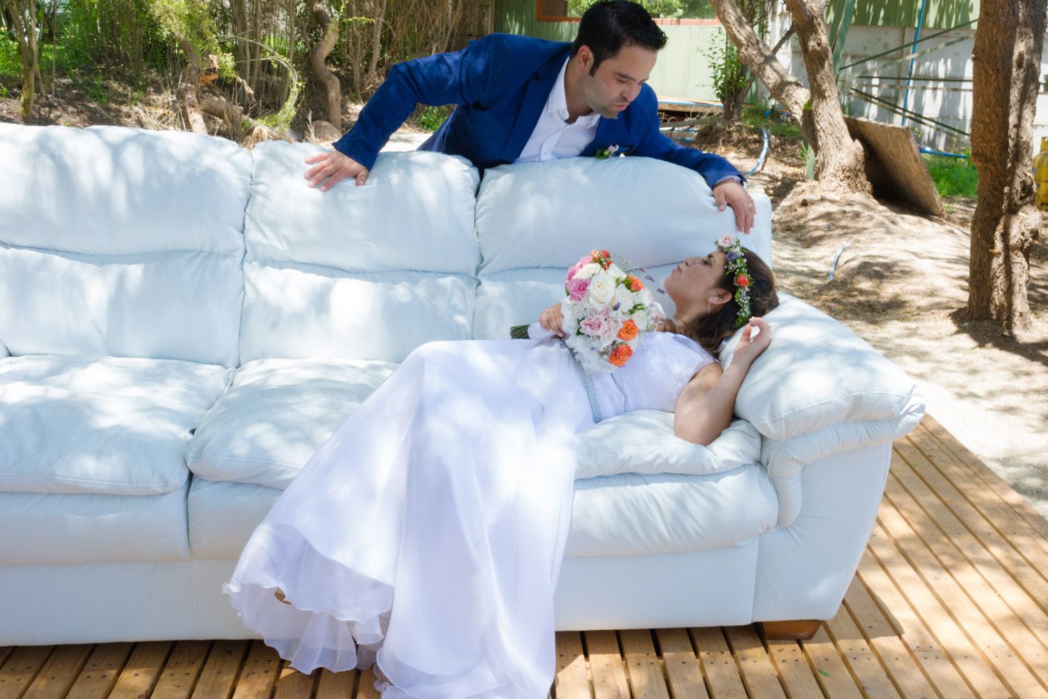 Groom in blue suit leans over bride lying on white outdoor sofa holding bridal bouquet.