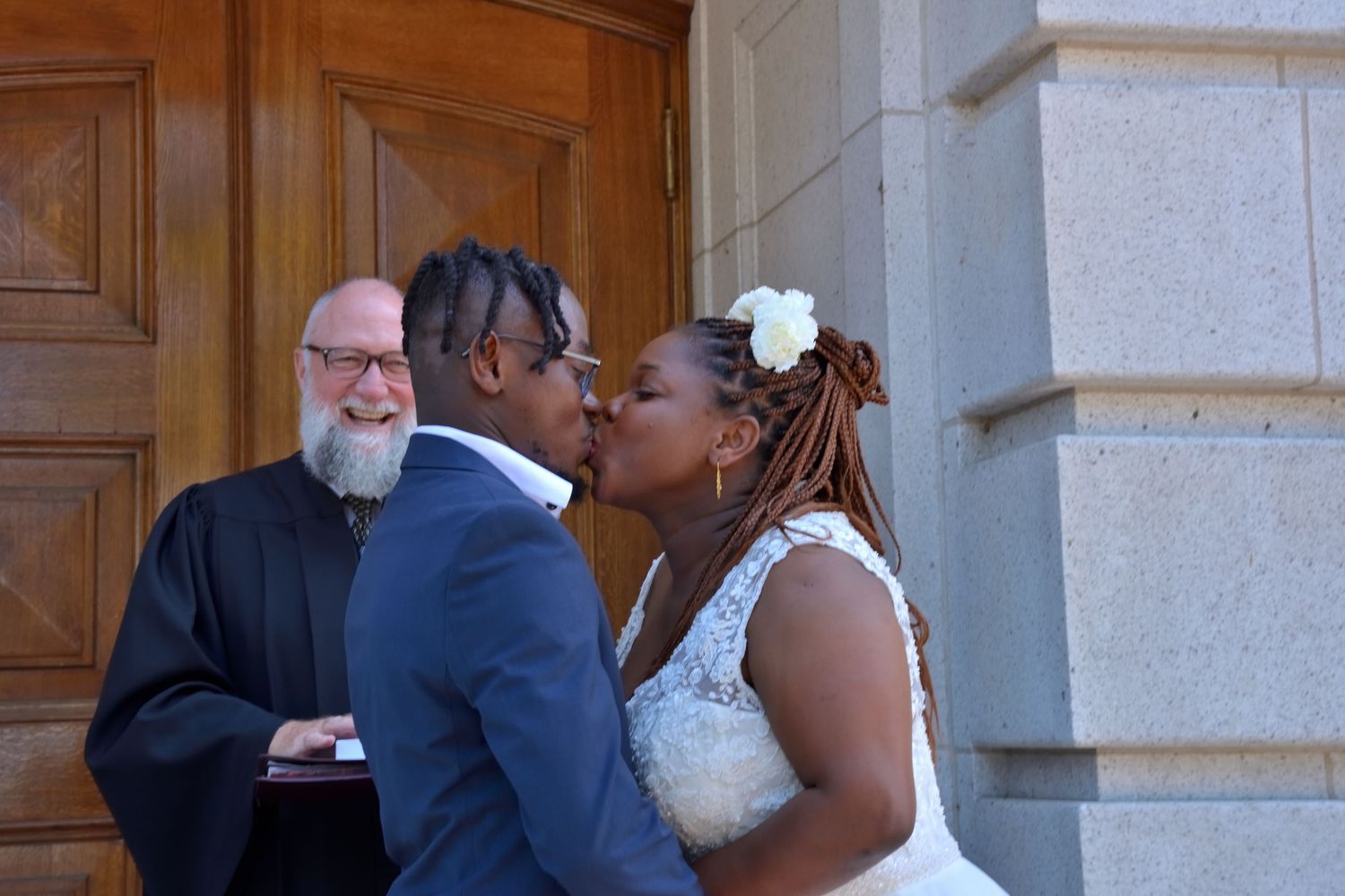 Newlyweds share first kiss as smiling officiant watches during outdoor wedding ceremony.