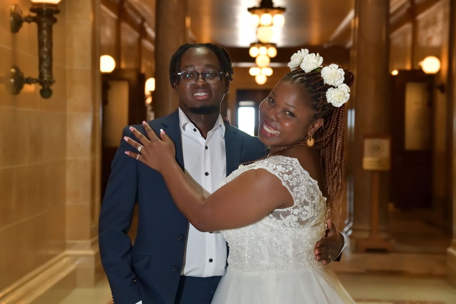 A smiling Black bride in white lace dress and groom in navy suit pose together in a grand hallway.