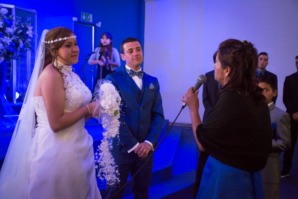 Bride and groom stand before officiant holding microphone during indoor wedding ceremony with blue lighting.