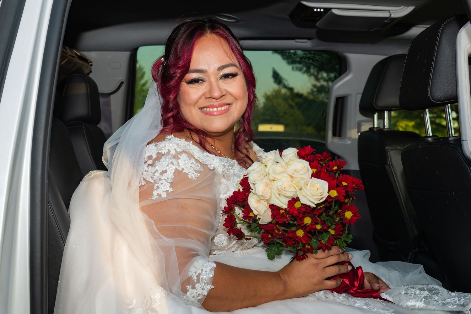 Smiling bride in lace wedding dress holding red and white bouquet seated in white limousine.
