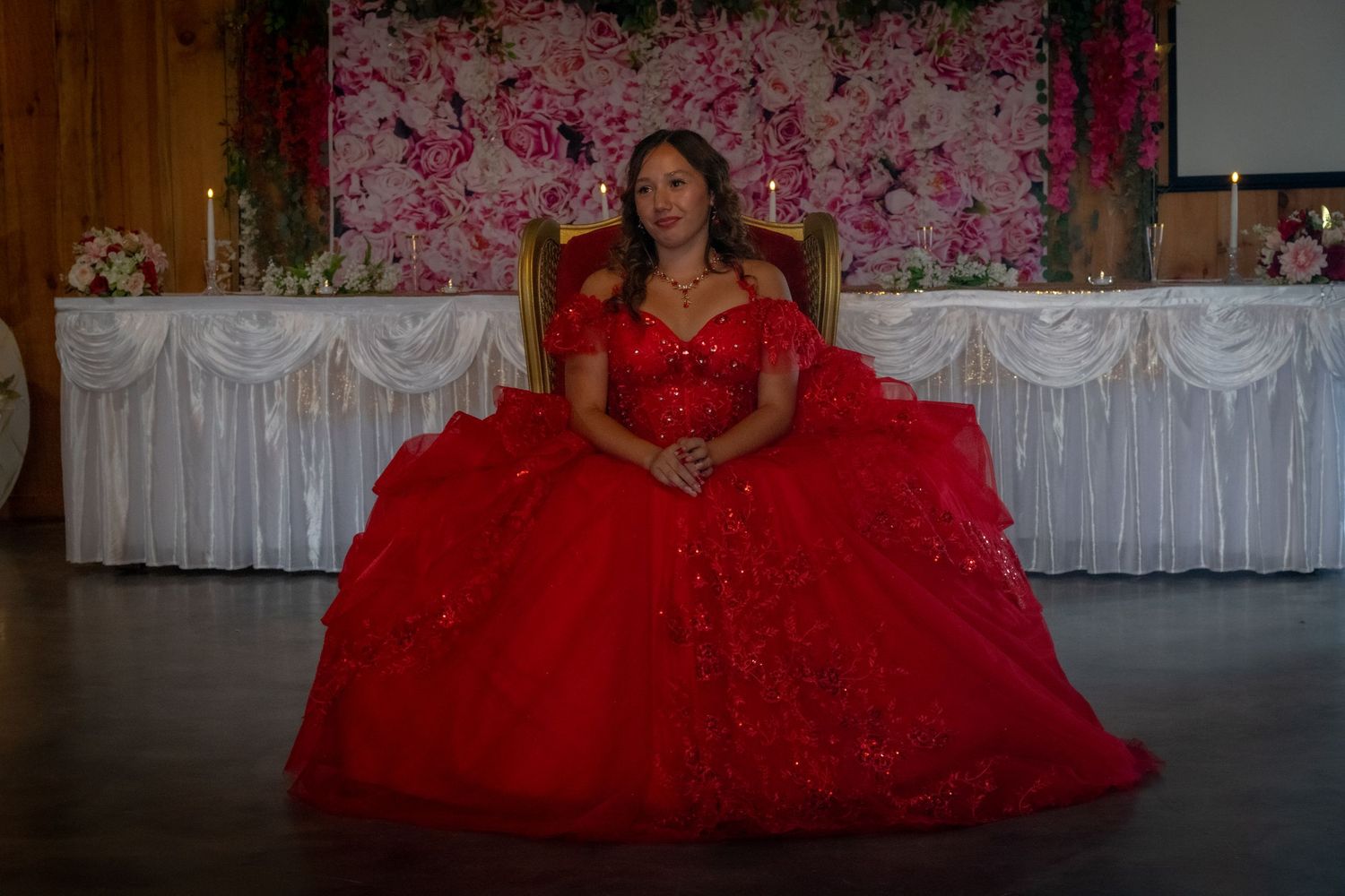A quinceañera girl sits regally on a golden throne in a sparkling red ball gown before a floral backdrop.