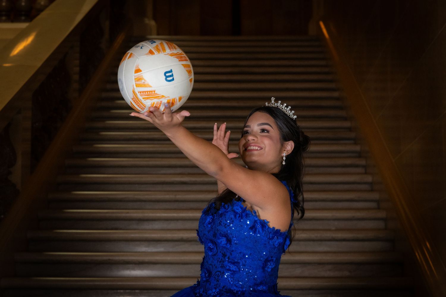 Young woman in blue quinceañera dress and tiara posing with volleyball on staircase.
