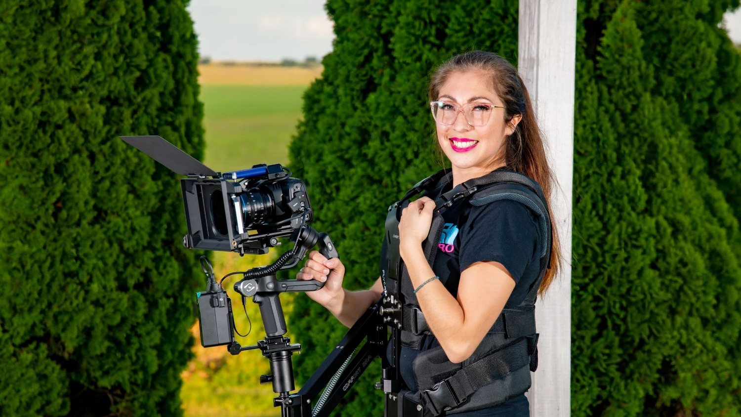 Bianca videographer smiling while holding a professional camera gimbal stabilizer outdoors near green trees.