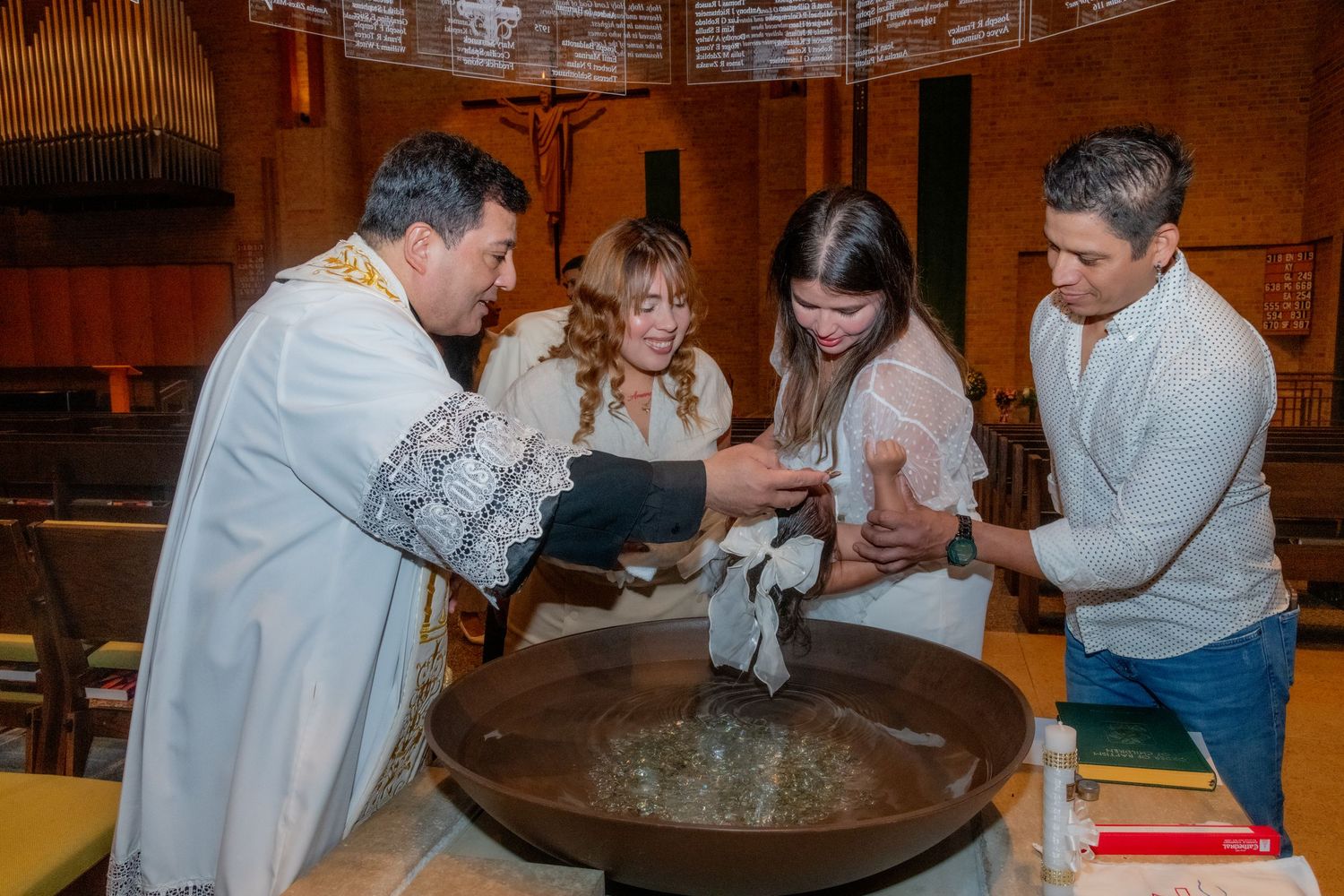 Catholic priest performing a baptism ceremony at a church font with family members present.