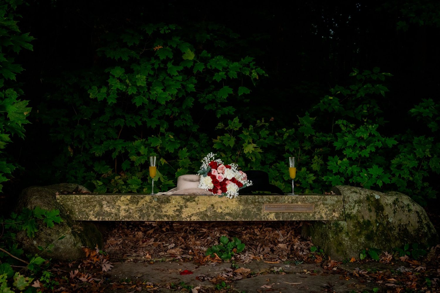 A wedding bouquet and champagne flutes sit on a stone bench surrounded by lush green foliage at night.