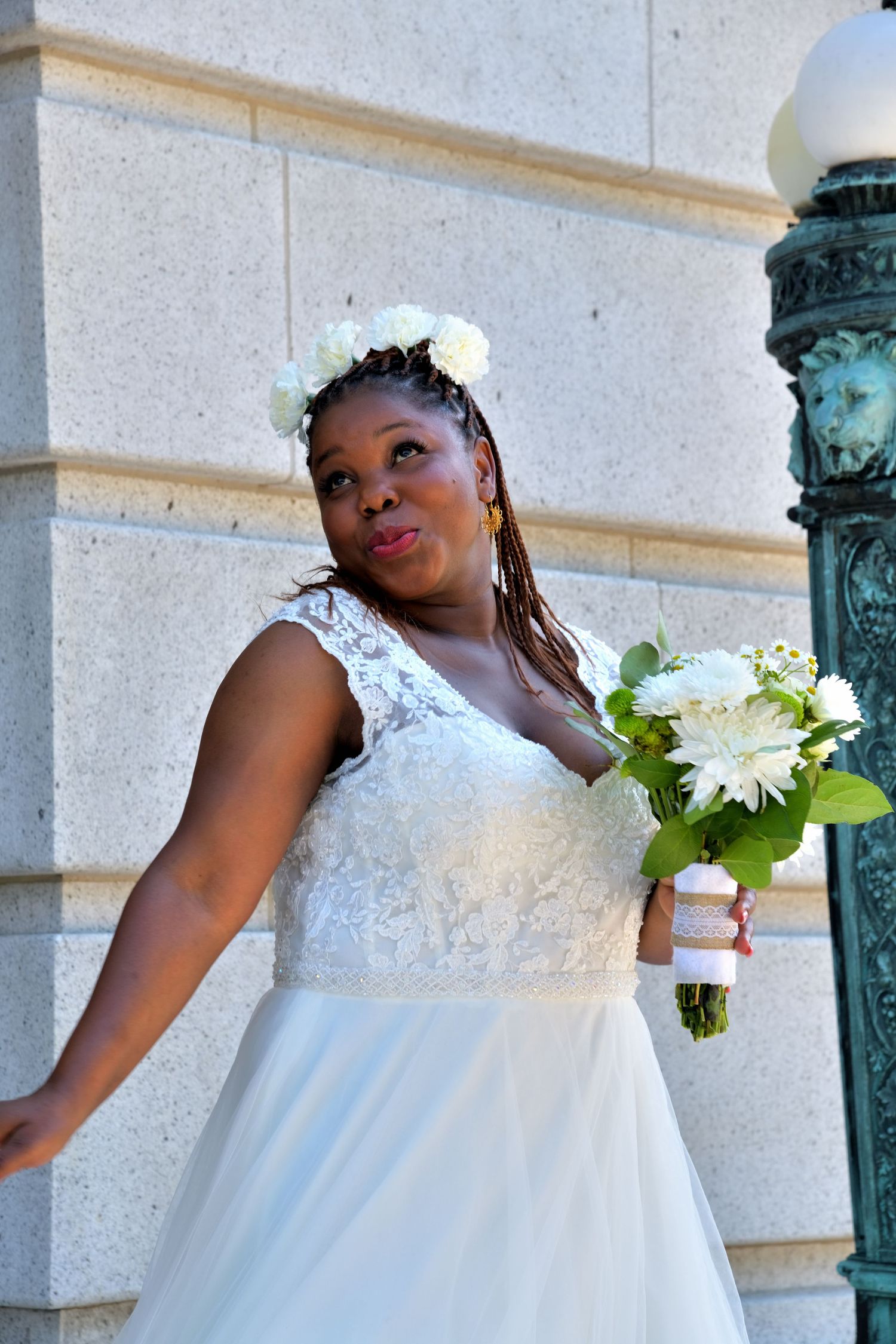 Bride in white lace gown with floral crown holds white bouquet beside stone column.