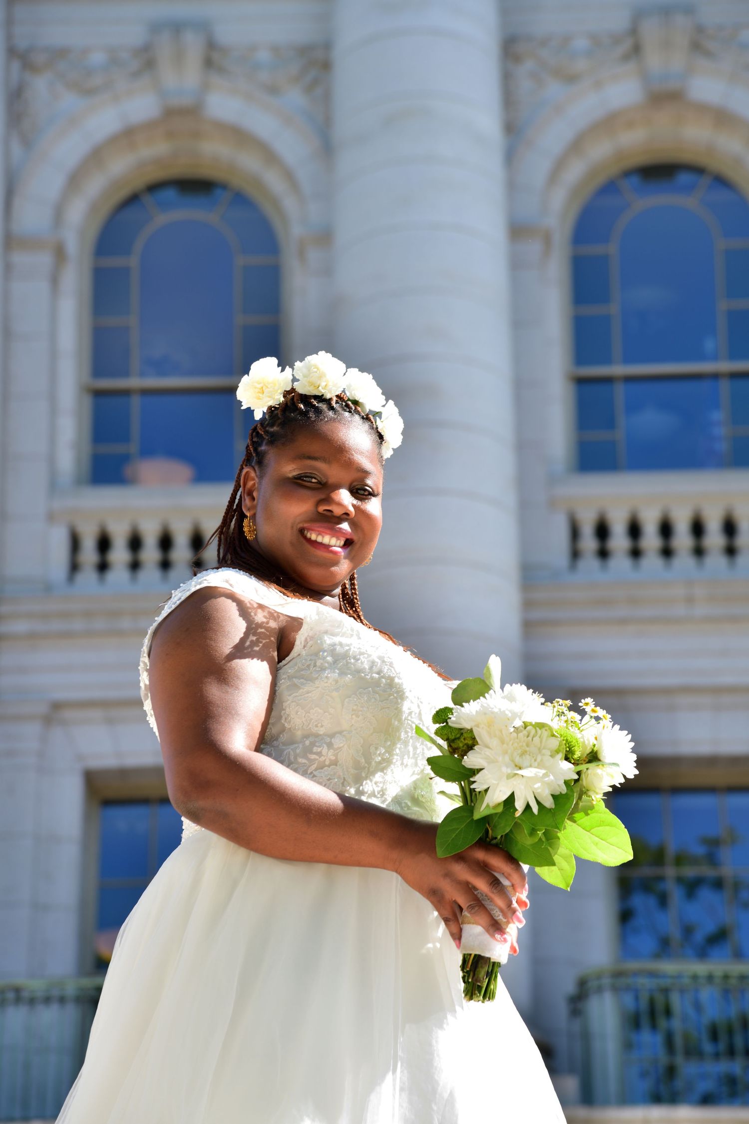 A radiant Black bride in a white ball gown holds a bouquet of white flowers outside a classical building.