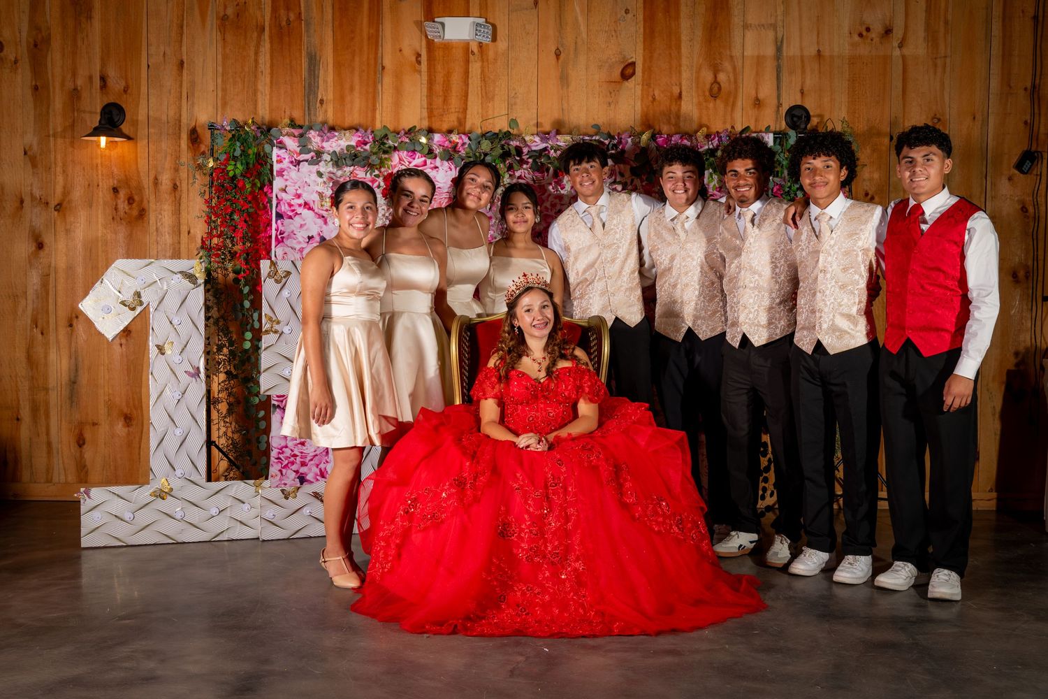 A quinceañera girl in a red ball gown poses with her court of honor in front of a floral photo backdrop.