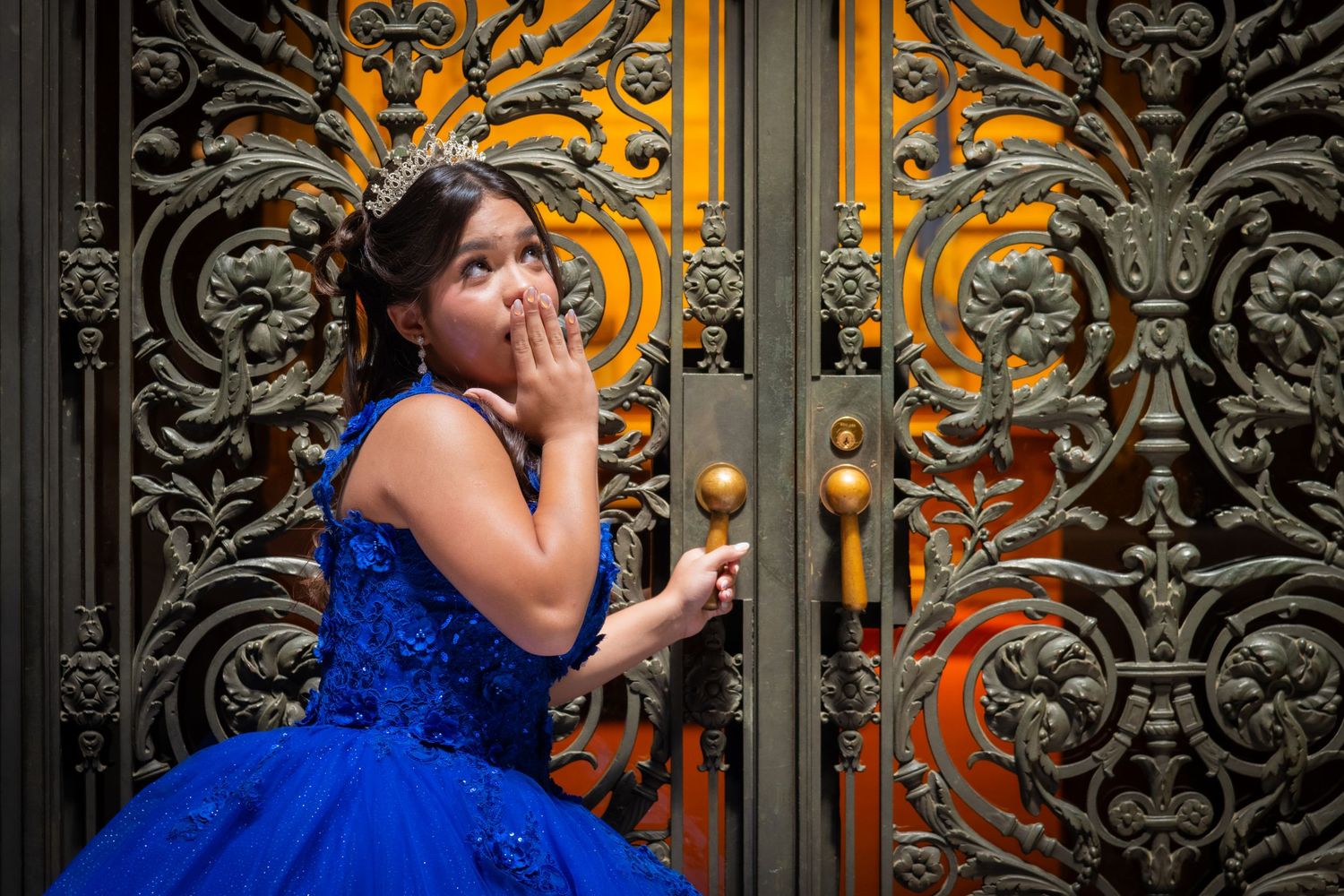 Young girl in royal blue quinceañera dress posing at ornate decorative iron doors.