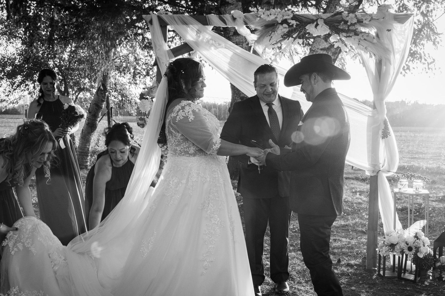Black and white photo of bride and groom exchanging vows under floral arch with officiant.