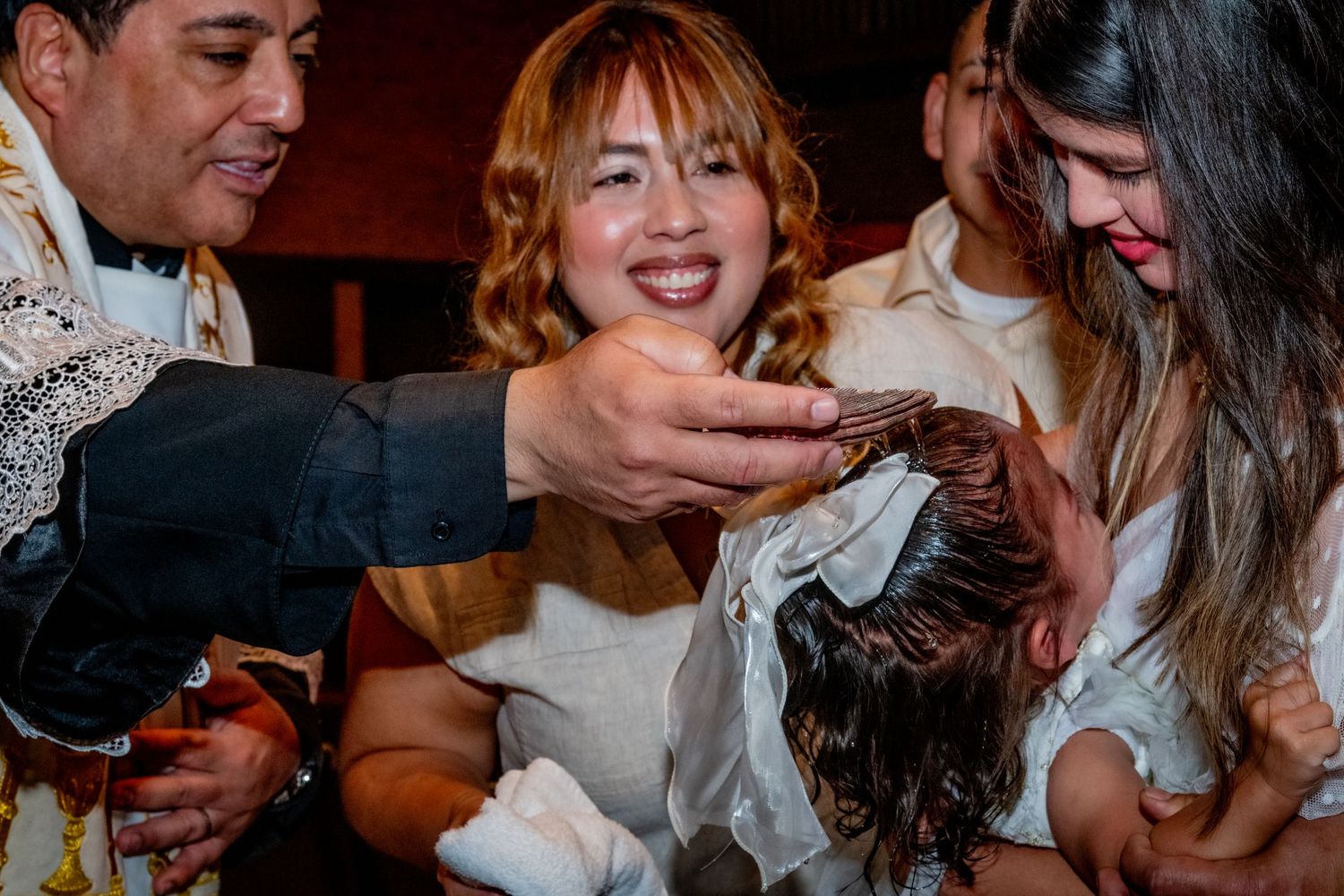 Priest performing a baptism ceremony, pouring water on a baby's head while smiling family members watch closely.