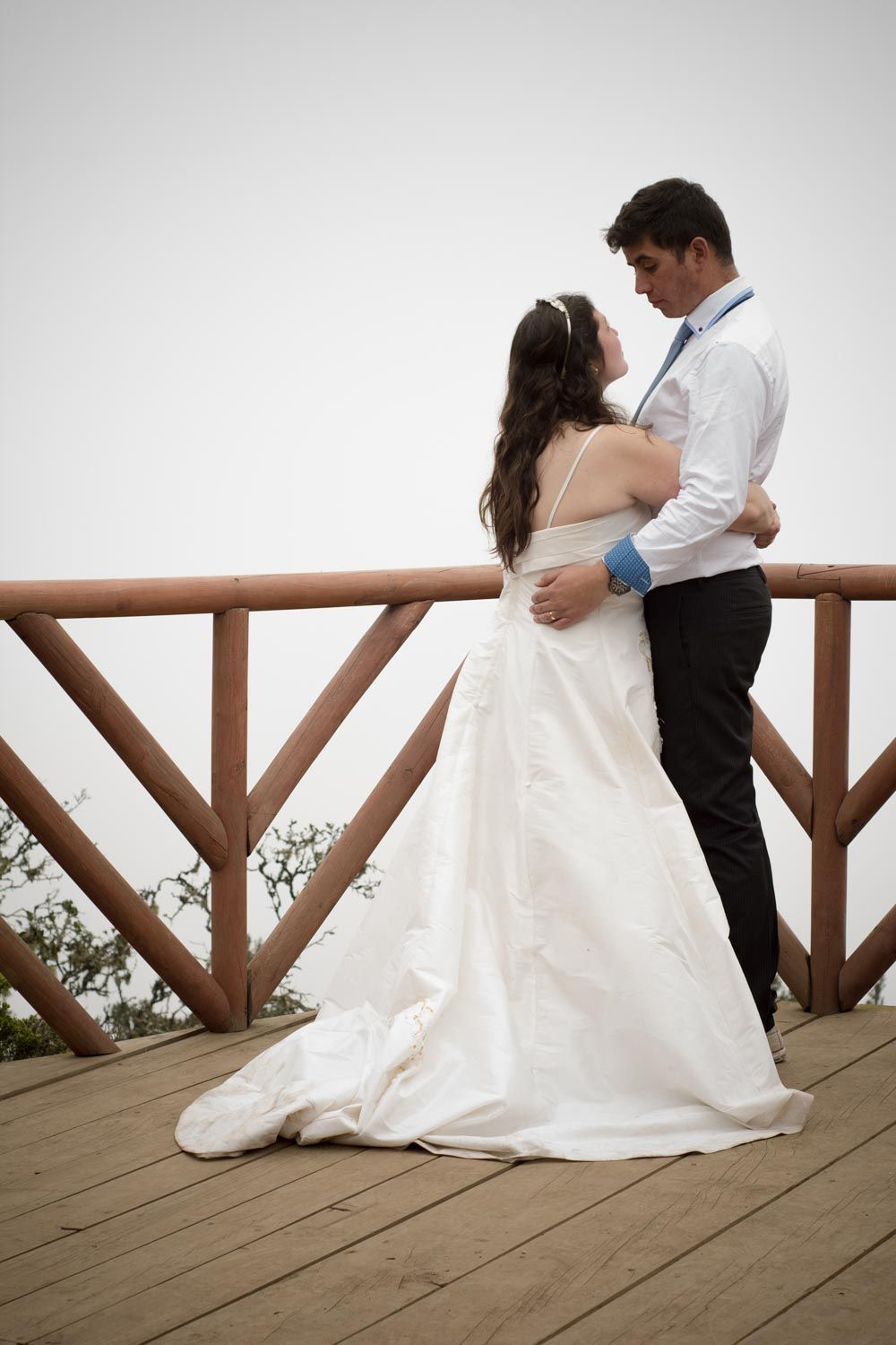 Bride and groom embracing romantically on wooden deck with open sky background.
