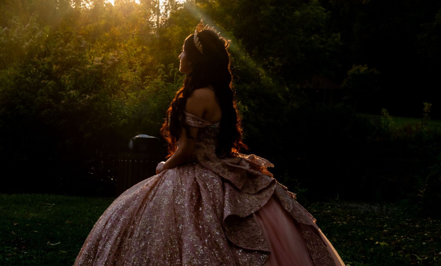 Silhouette of quinceañera girl in rose gold ballgown in golden sunlight among lush green trees.