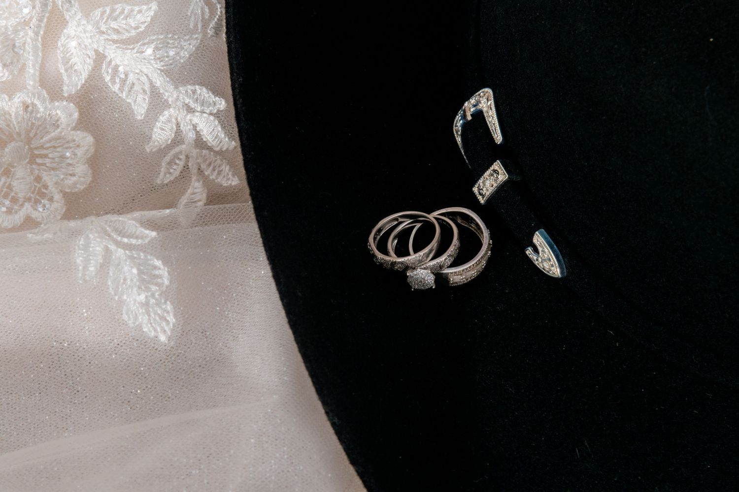 Wedding rings and diamond earrings arranged elegantly on a black velvet hat beside white lace fabric.