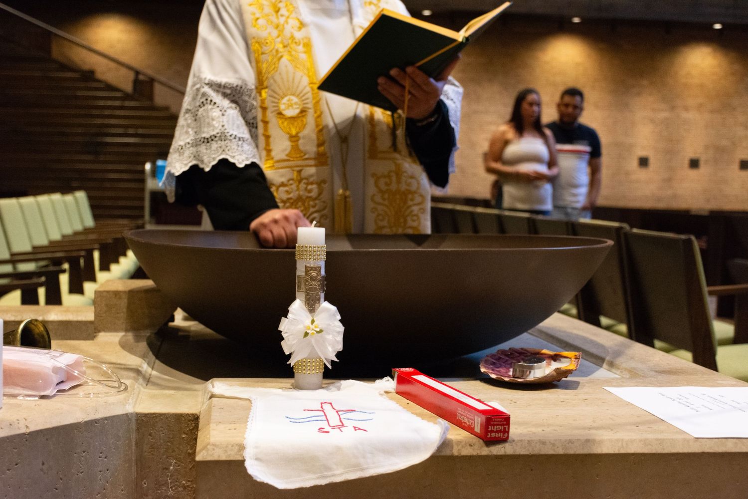 Priest performing a baptism ceremony at a church font with candle and white cloth in the foreground.